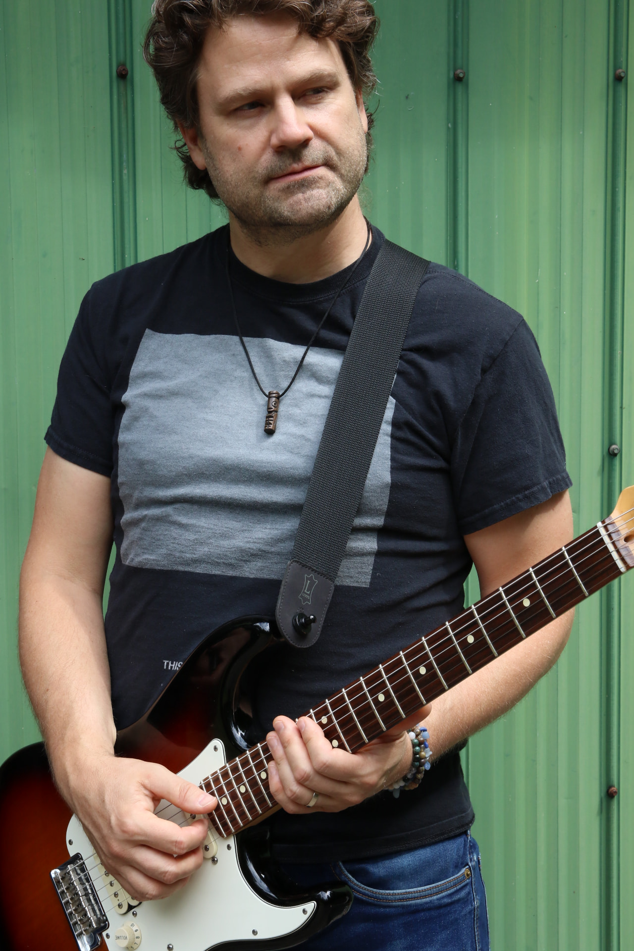 Kevin holding a sunburst Stratocaster in front of a green corrugated wall